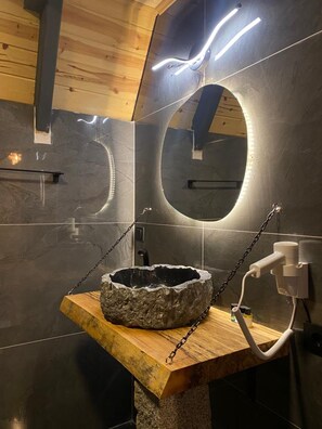 A modern bathroom with a stone sink on a wooden platform, surrounded by dark tiles and illuminated by under-cabinet lighting. - Berva Bungalow (Çamlıhemşin)