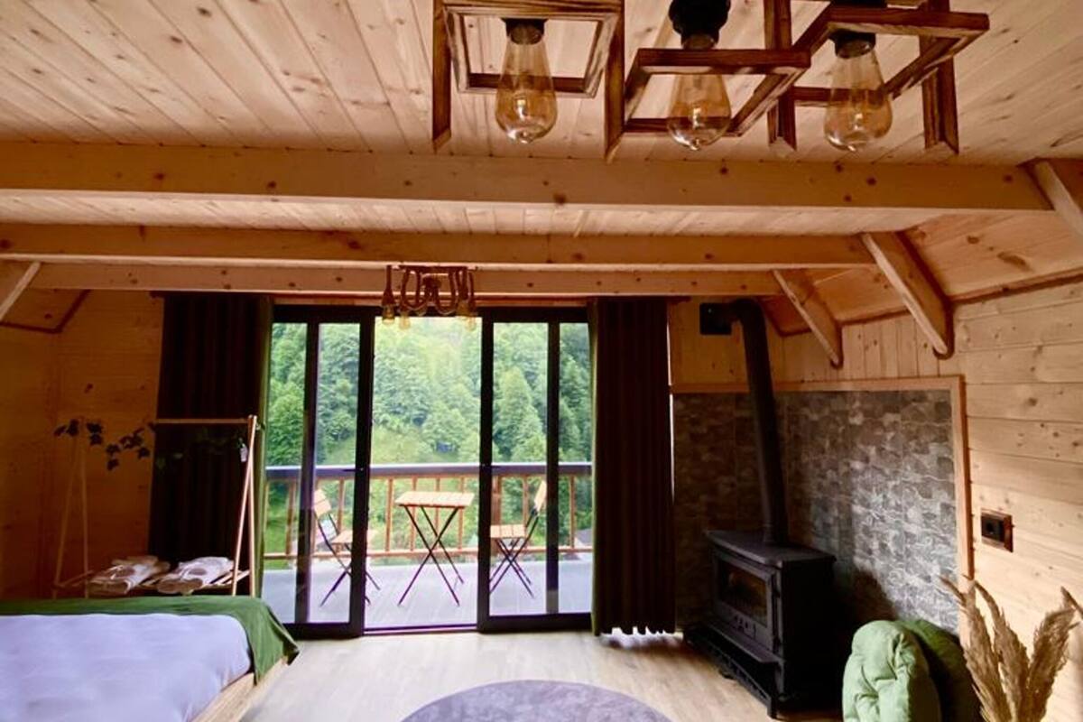 Ein Holzhütten-Interieur mit einem Bett, einem runden Teppich, einem Kamin und einem Balkon mit Blick auf Grünfläche.