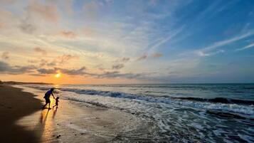Escena de playa con atardecer, olas y una persona caminando sobre la arena.