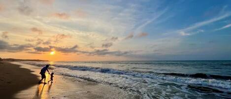 Escena de playa con atardecer, olas y una persona caminando sobre la arena.