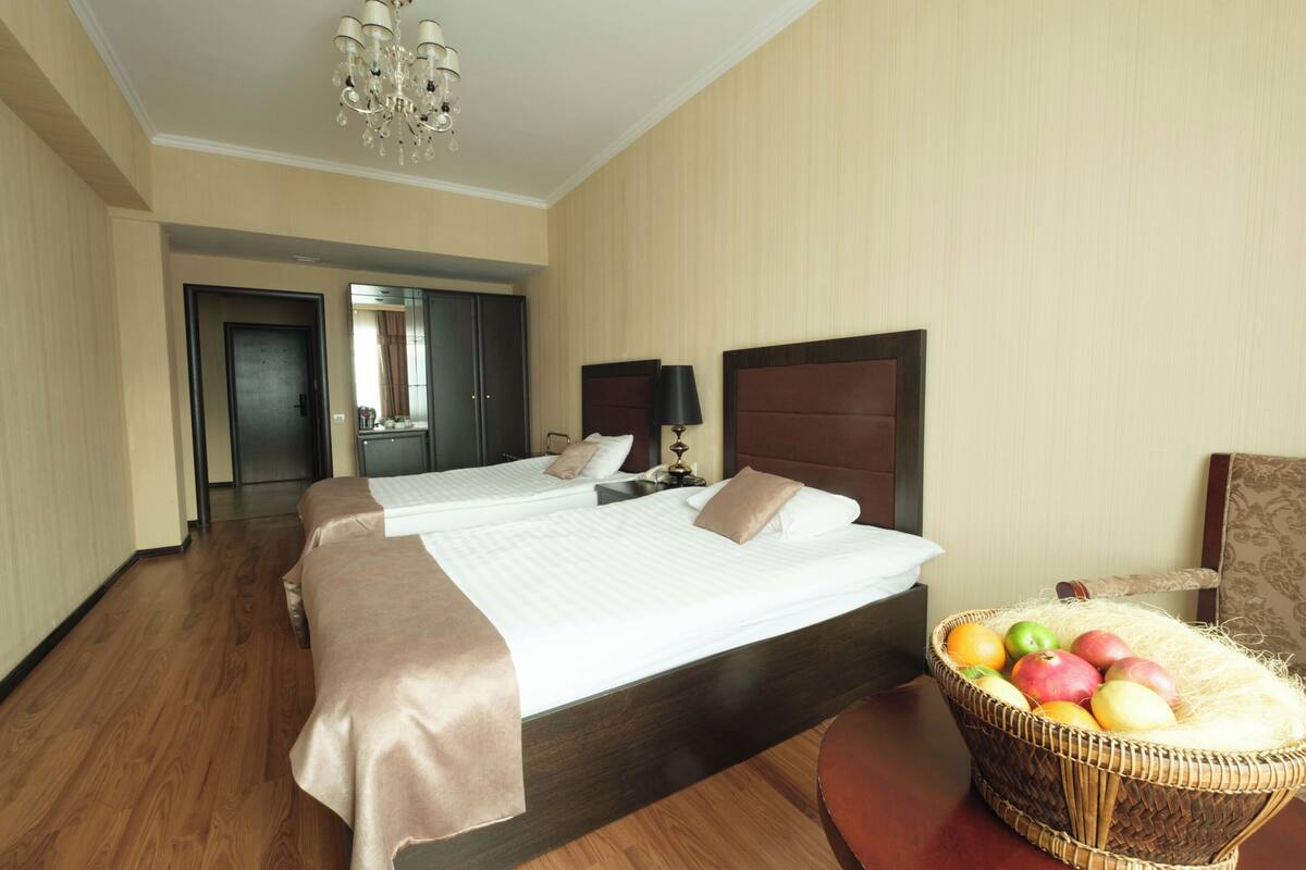 Hotellihuone, jossa on kaksi sänkyä, kattokruunu, peili, yöpöytä lampun kanssa ja hedelmäkori.