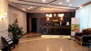 A hotel reception area with a reception desk, a potted plant, a sofa, and a wall-mounted clock.