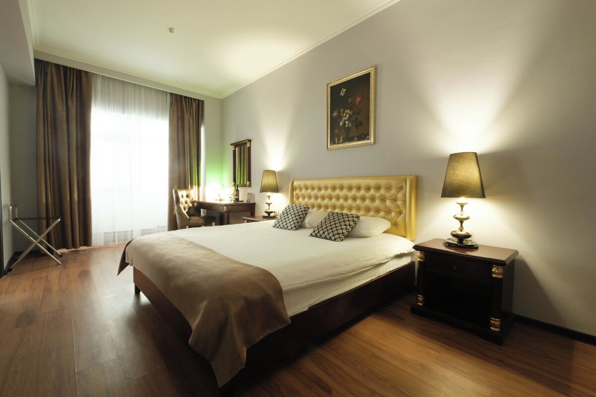 A hotel room with a large bed, two bedside lamps, a painting on the wall, and a desk with a chair.