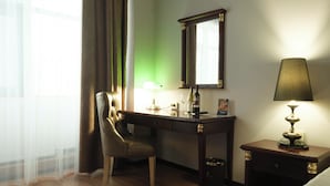 A hotel room with a wooden desk, a chair, a lamp, a mirror, and a bedside table.