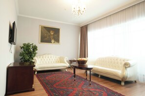 A living room with two white sofas, a wooden coffee table, a red patterned rug, a potted plant, a framed picture on the wall, and a chandelier. - Reikartz Astana (Astana)