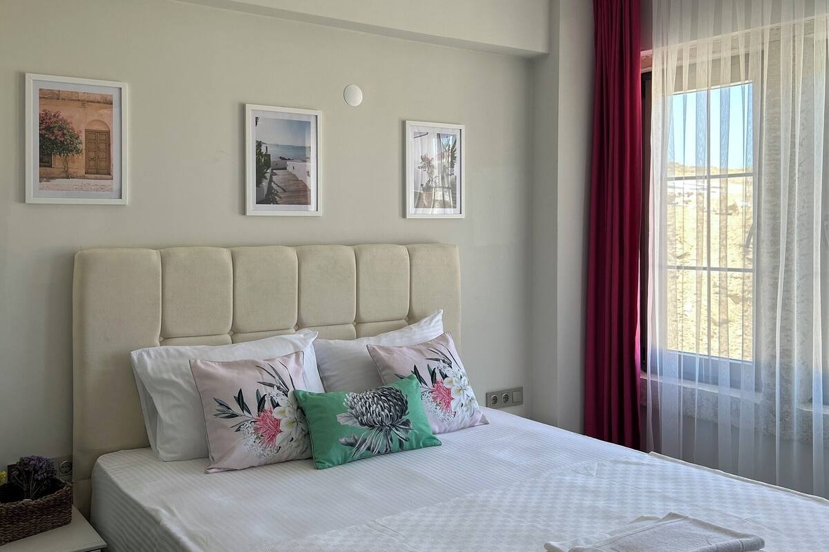 Ein ordentlich bezogenes Bett mit Kissen, ein Nachttisch mit einem Korb und ein Fenster mit Vorhängen.