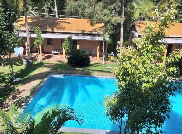 A swimming pool surrounded by greenery and a building with a tiled roof. - Nikhara Garden And Resorts (Mandya)