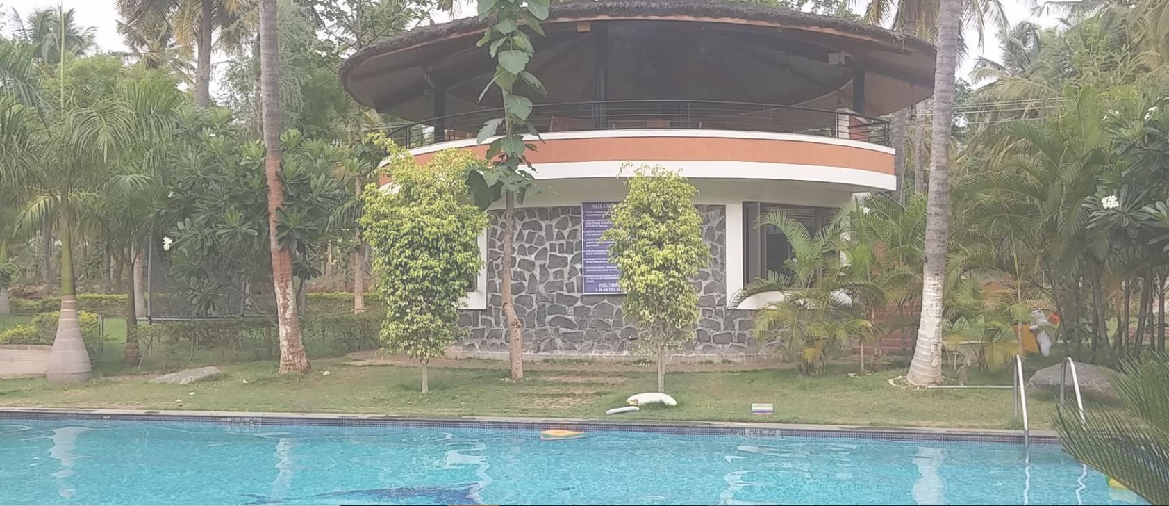Vista de una piscina frente a un edificio con fachada de piedra, un balcón circular y vegetación tropical.