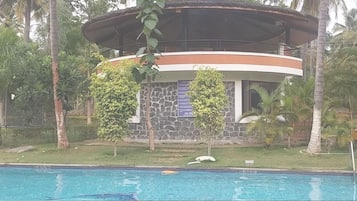 Vista de una piscina frente a un edificio con fachada de piedra, un balcón circular y vegetación tropical.