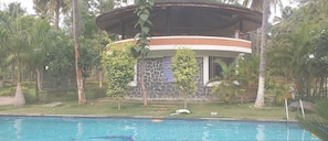 A poolside view of a building with a stone facade, a circular balcony, and tropical vegetation. - Nikhara Garden And Resorts (Mandya)