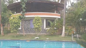 Vue sur un bâtiment avec une façade en pierre, un balcon circulaire et une végétation tropicale, le tout vu depuis la piscine.
