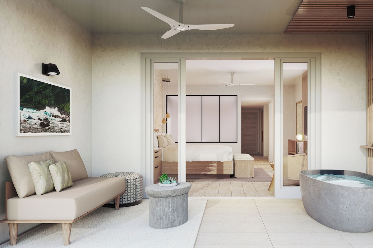 A modern bedroom with a bed, a sitting area with a sofa, a large bathtub, and a ceiling fan.