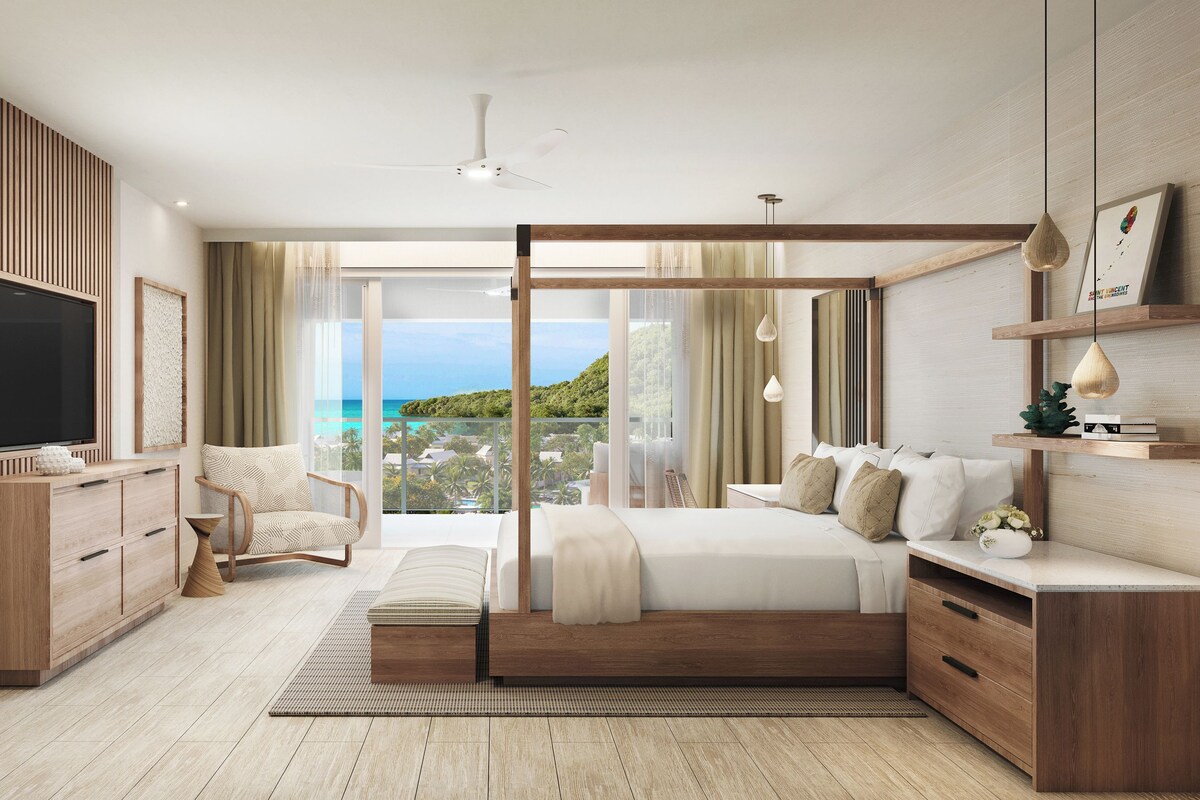 A modern bedroom with a large bed, a TV, a chair, and a view of the ocean.