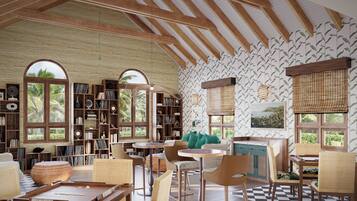 Ein Zimmer mit hohen Decken, Holzbalken und großen Fenstern mit tropischem Wandpapier. Es gibt Bücherregale, Korbmöbel und einen Blick auf Palmen.