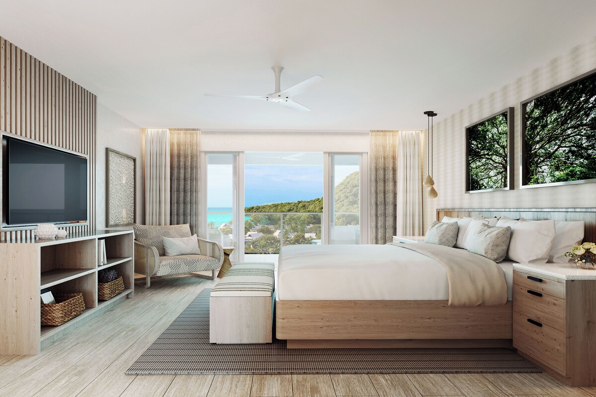 A bedroom with a bed, a TV, a wooden headboard, a large window with a view, and a ceiling fan.