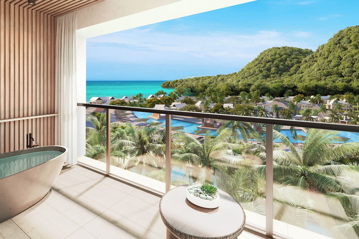 Casa de banho moderna com uma banheira independente, uma mesinha redonda com uma planta, e uma varanda com vista para uma piscina e paisagem tropical.