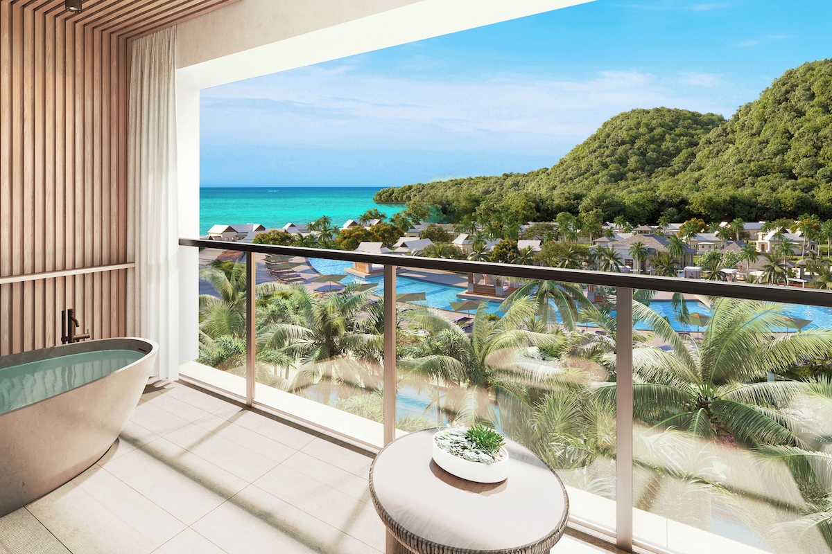 A modern bathroom with a freestanding bathtub, a small round table with a plant, and a balcony with a view of a pool and tropical landscape.