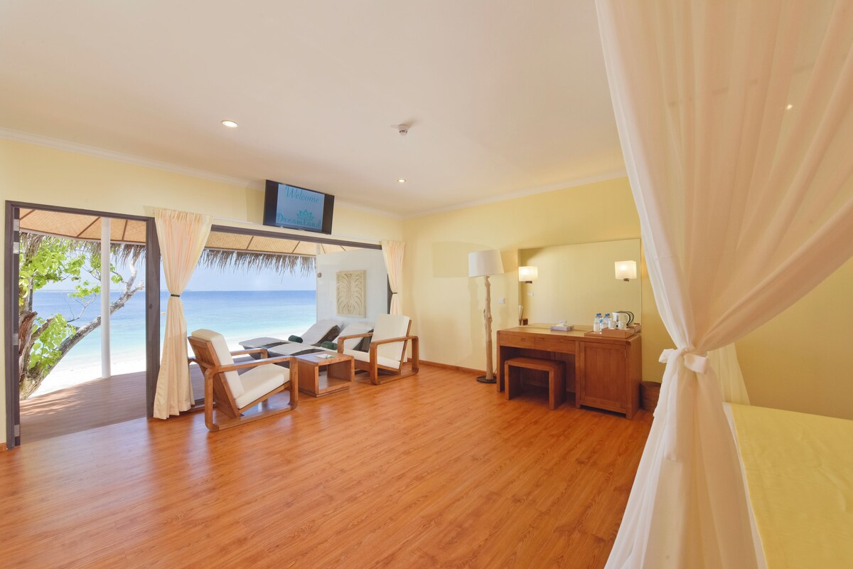 A room with a wooden floor, a desk with a lamp, a chair, and a bed. There is a TV on the wall and a view of the ocean through a glass door.