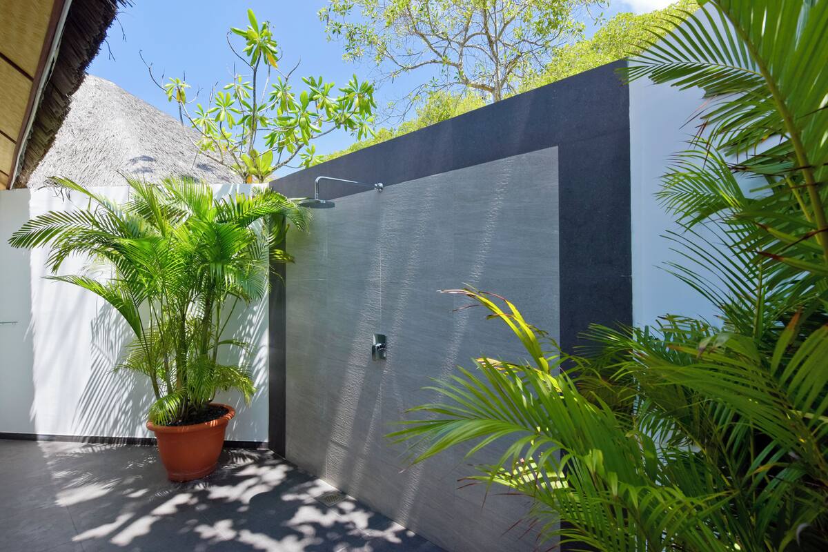 A modern outdoor shower area with a concrete wall, a potted plant, and a thatched roof structure in the background.