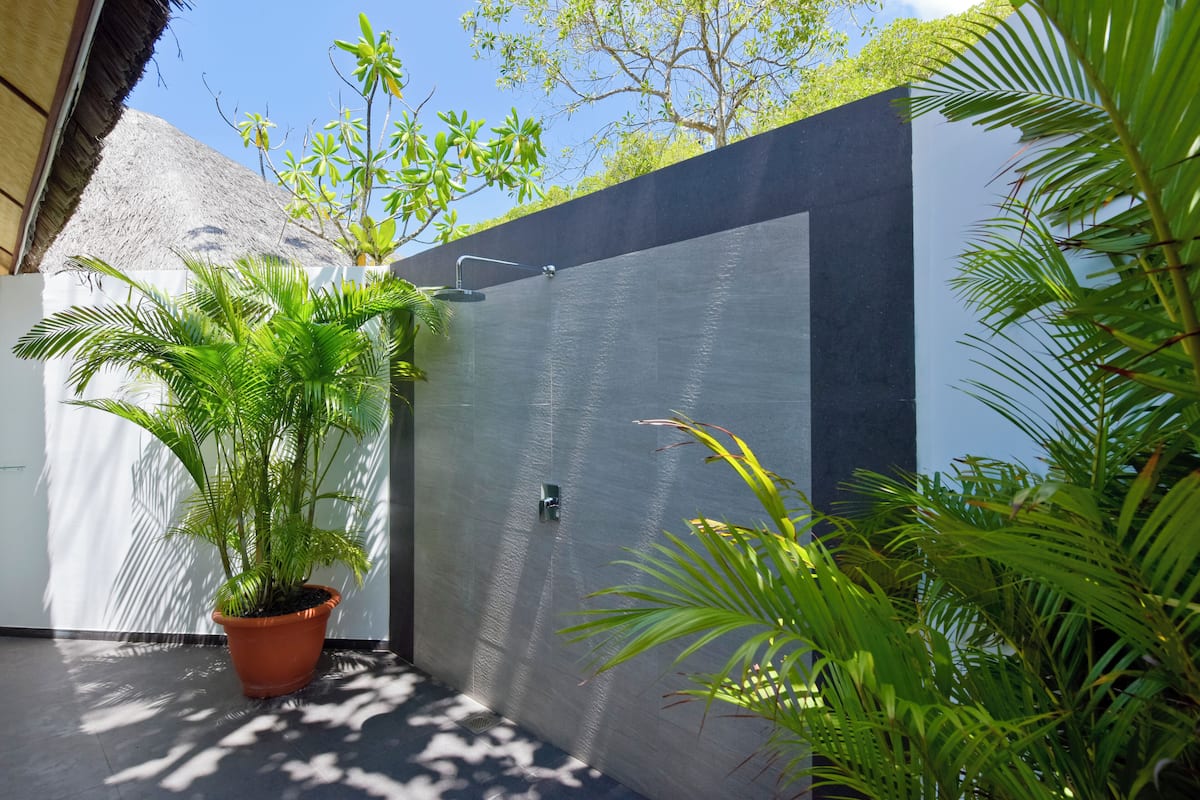 Un espace de douche extérieur moderne, avec un mur en béton, une plante en pot et, au fond, une structure dotée d’un toit en chaume.