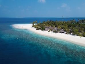 An aerial view of a tropical island with a white sandy beach, clear turquoise waters, and thatched-roof huts.