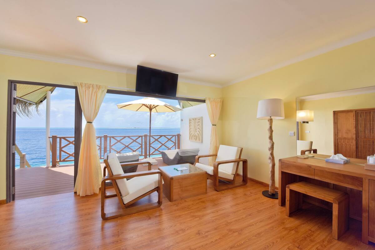 A spacious room with wooden flooring, a large window with a view of the sea, a wooden desk, a chair, a sofa, and a lamp.