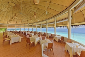 Uma ampla área de refeições interna, com mesas e cadeiras, grandes janelas com vista para o mar e teto alto com luminárias pendentes.