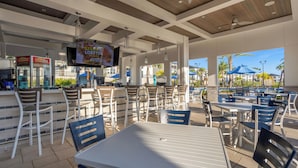 An outdoor dining area with tables and chairs, a bar area with a television, and a view of palm trees and a pool. - Champions Gate Resort Homes (Davenport)