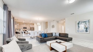 A modern living room with a sectional sofa, a white coffee table, and a dining area in the background. - Champions Gate Resort Homes (Davenport)