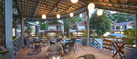 Un patio exterior con suelo de madera, mobiliario de mimbre y lámparas colgantes.