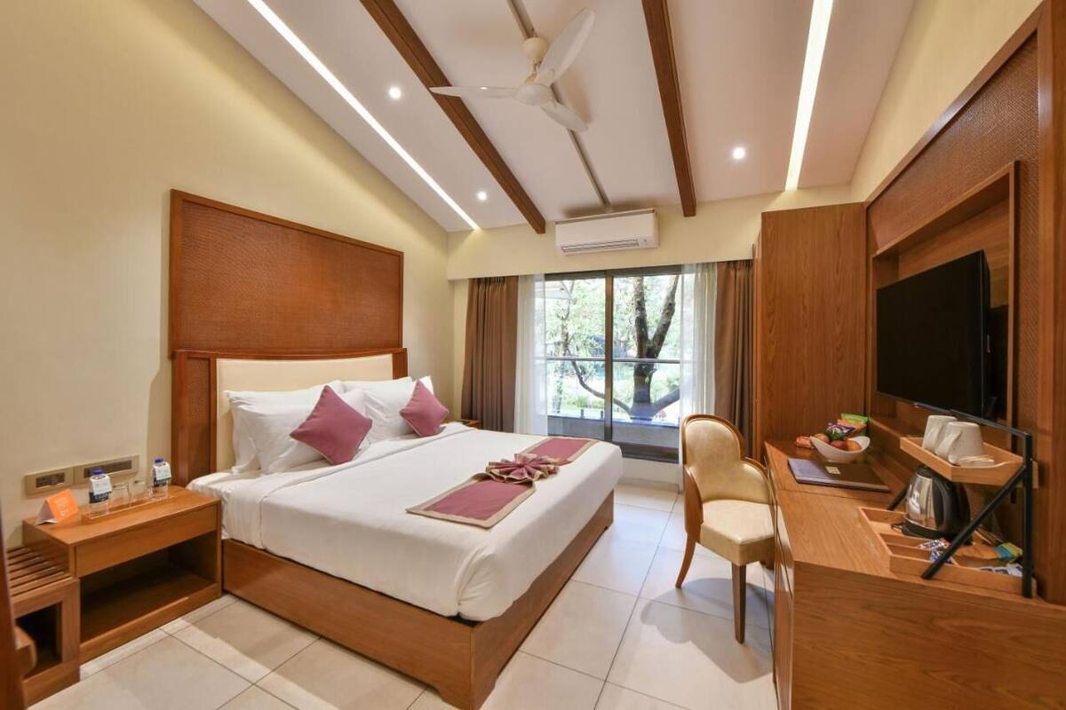 Habitación de hotel con una cama grande, un escritorio con televisor, una silla y una ventana con vistas a árboles.