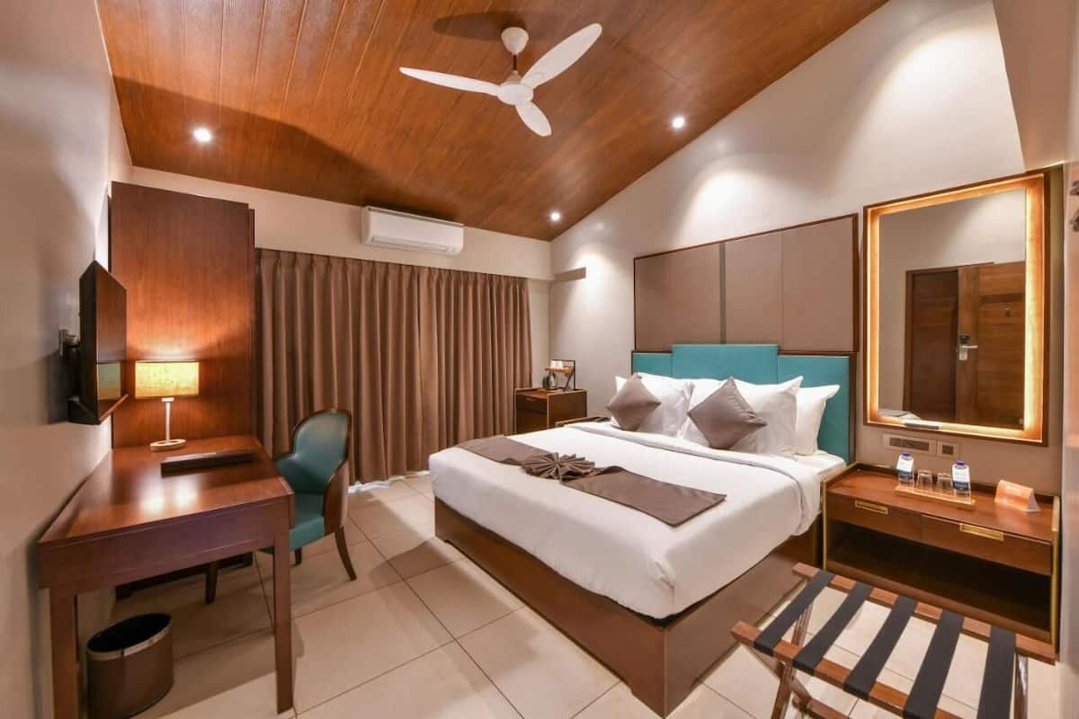 Habitación de hotel con cama, escritorio, silla, mesita de noche, espejo, ventilador de techo y cortinas.