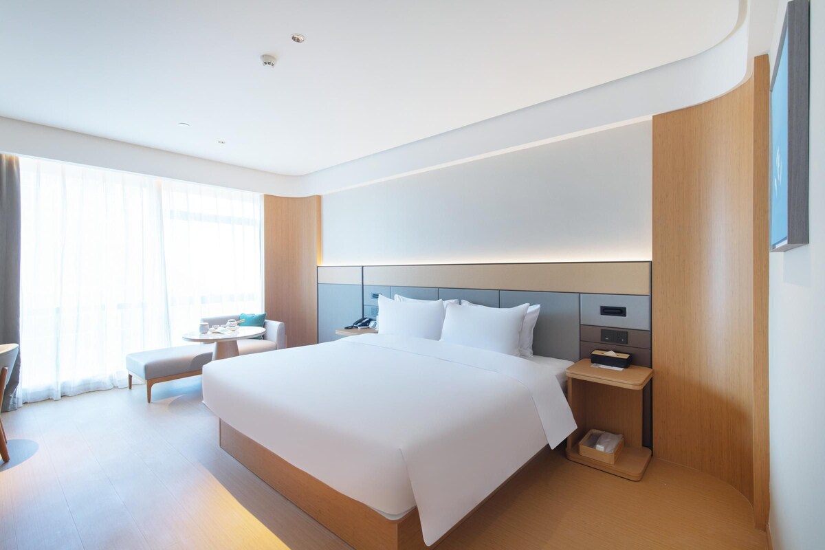 A modern hotel room with a large bed, bedside tables, a small seating area, and a large window.