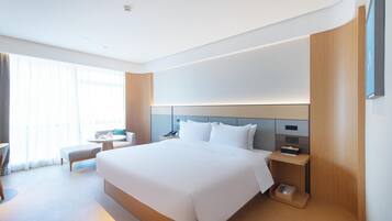 Una habitación de hotel moderna con una cama grande, mesitas de noche, un pequeño rincón de descanso y un amplio ventanal.