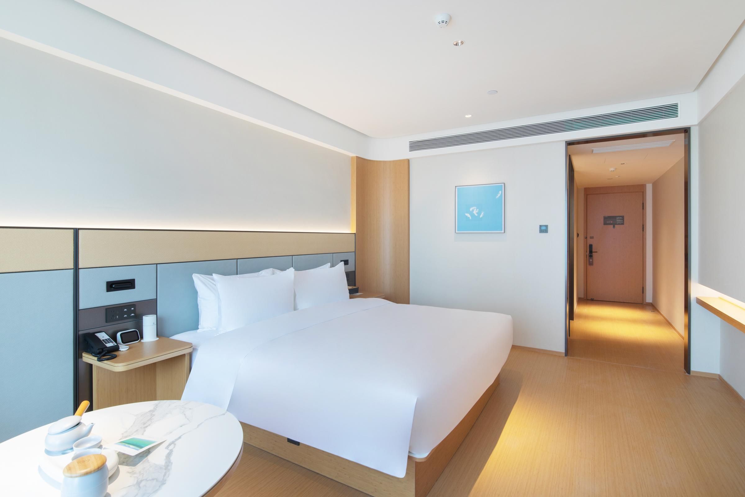 A modern hotel room with a large bed, bedside table, and a small round table with a cup and a book.