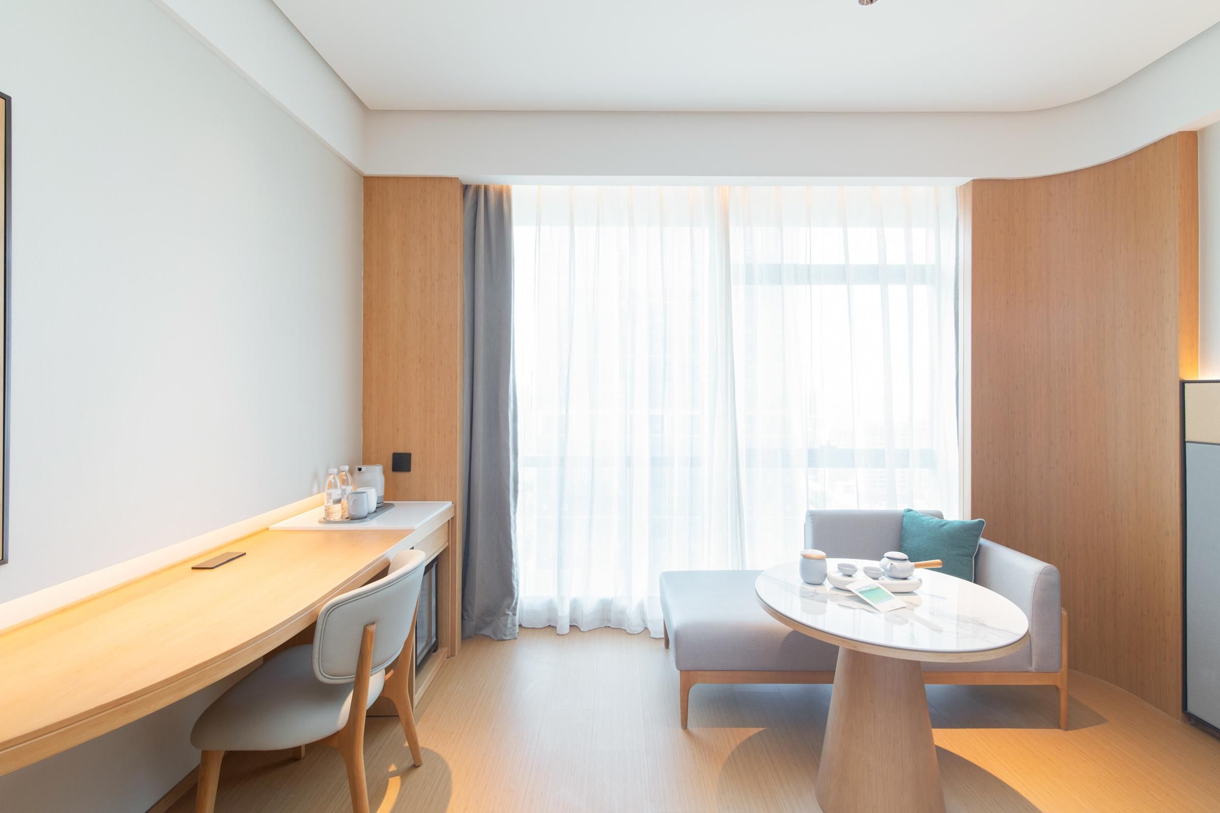 Una habitación de hotel moderna con un escritorio de madera, una mesa redonda, un sofá y un ventanal con cortinas traslúcidas.