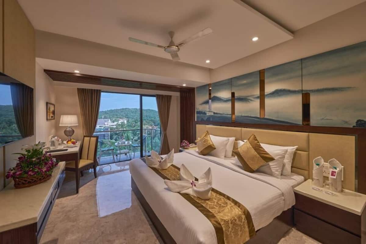 A hotel room with a large bed, a desk, a chair, a TV, and a balcony with a view.