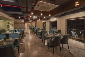 A restaurant with a long dining area, multiple tables, and chairs. There are hanging lights and a large window on one side. - Regenta Central Imperial Candolim (Candolim)