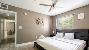 Un dormitorio con cama, ventilador de techo, ventana con persianas y una obra de arte colgada en la pared.