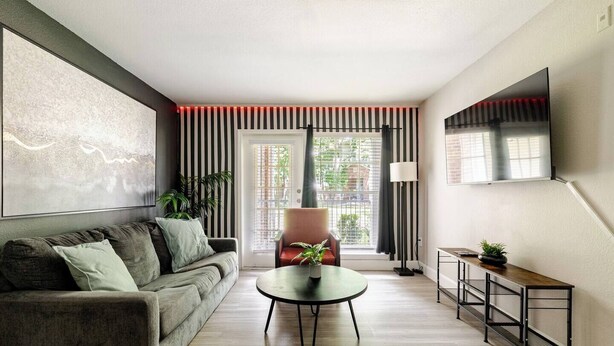 Una sala de estar moderna con sofá, una mesa de centro, una silla, un televisor montado en la pared y una ventana con persianas.