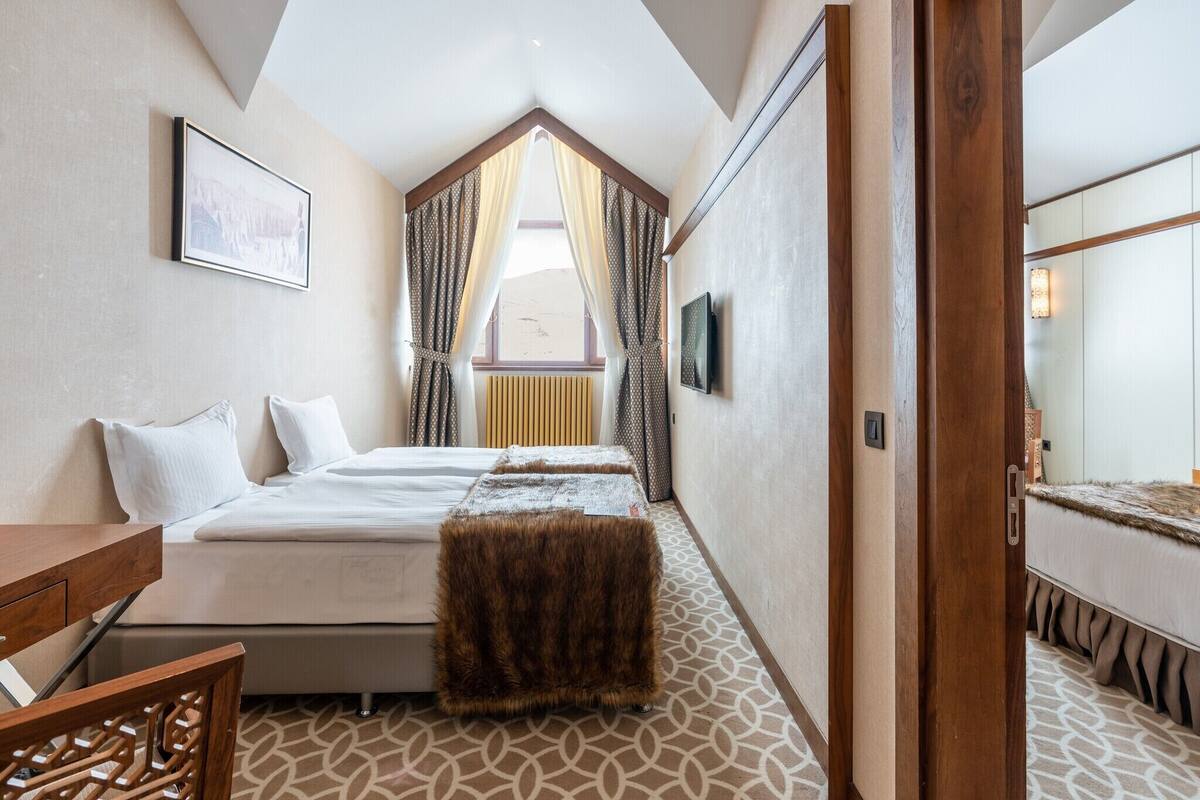 Ein Schlafzimmer mit einem Bett, einem Holztisch, einem Fellhocker, einem Fenster mit Vorhängen, einem gerahmten Bild an der Wand und einem gemusterten Boden.