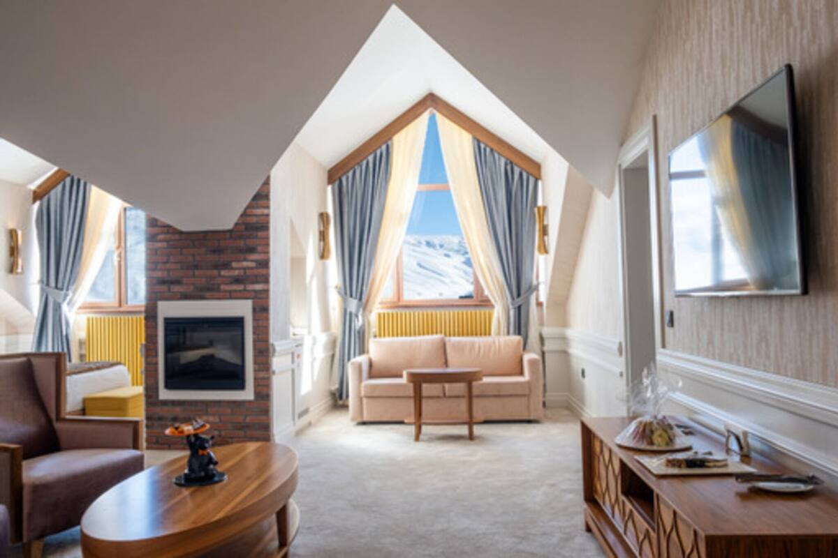 Ein modernes Hotelzimmer mit schrägverdeckter Decke, einem großen Fenster mit Vorhängen, einem Flachbildfernseher, einem Sofa, einem Couchtisch und einem Kamin.