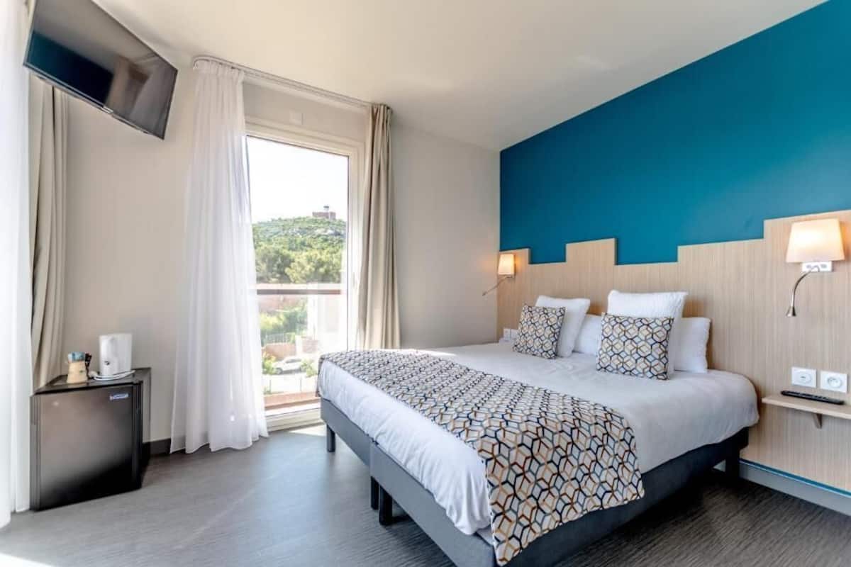 A hotel room with a large bed, a TV mounted on the wall, a small bedside table, and a window with curtains.