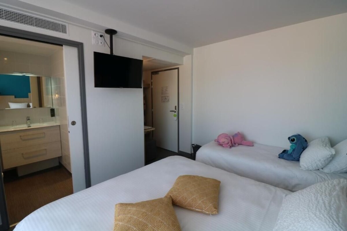 A hotel room with two beds, a TV mounted on the wall, and a bathroom visible through an open door.