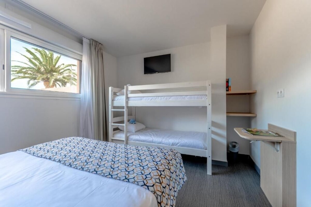 A hotel room with a bunk bed, a single bed, a TV on the wall, a small desk with a magazine, and a window with a view of palm trees.