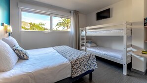A hotel room with a bed, a bunk bed, a desk, a TV, and a window with a view of palm trees.