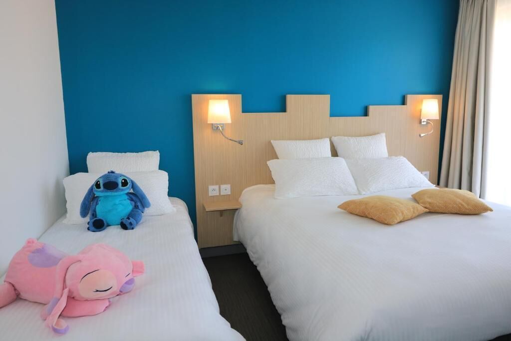 Two hotel beds with white bedding, a blue wall, and wall-mounted lamps.