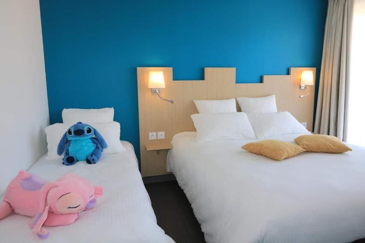 Two hotel beds with white bedding, a blue wall, and wall-mounted lamps.