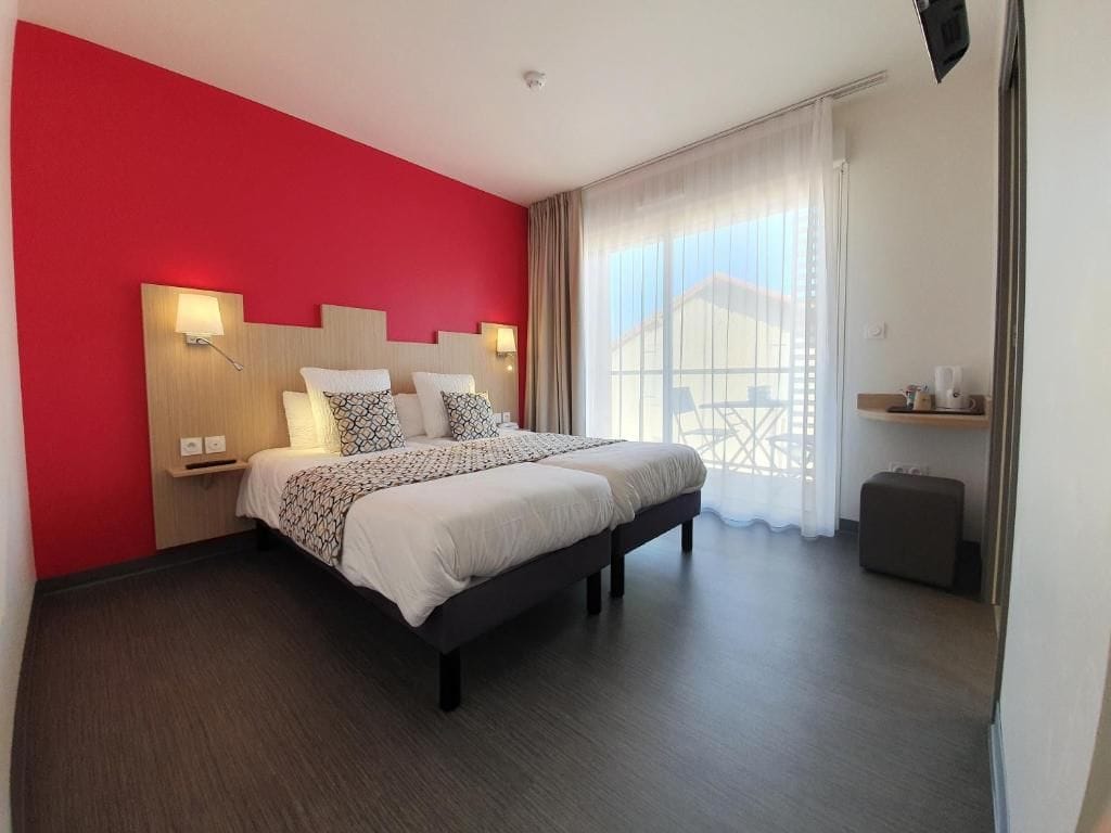 Una camera d'hotel moderna con un grande letto, una parete d'accento rossa e vista su un edificio attraverso tende trasparenti.