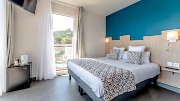A hotel room with a large bed, a TV mounted on the wall, a small bedside table, and a window with curtains.
